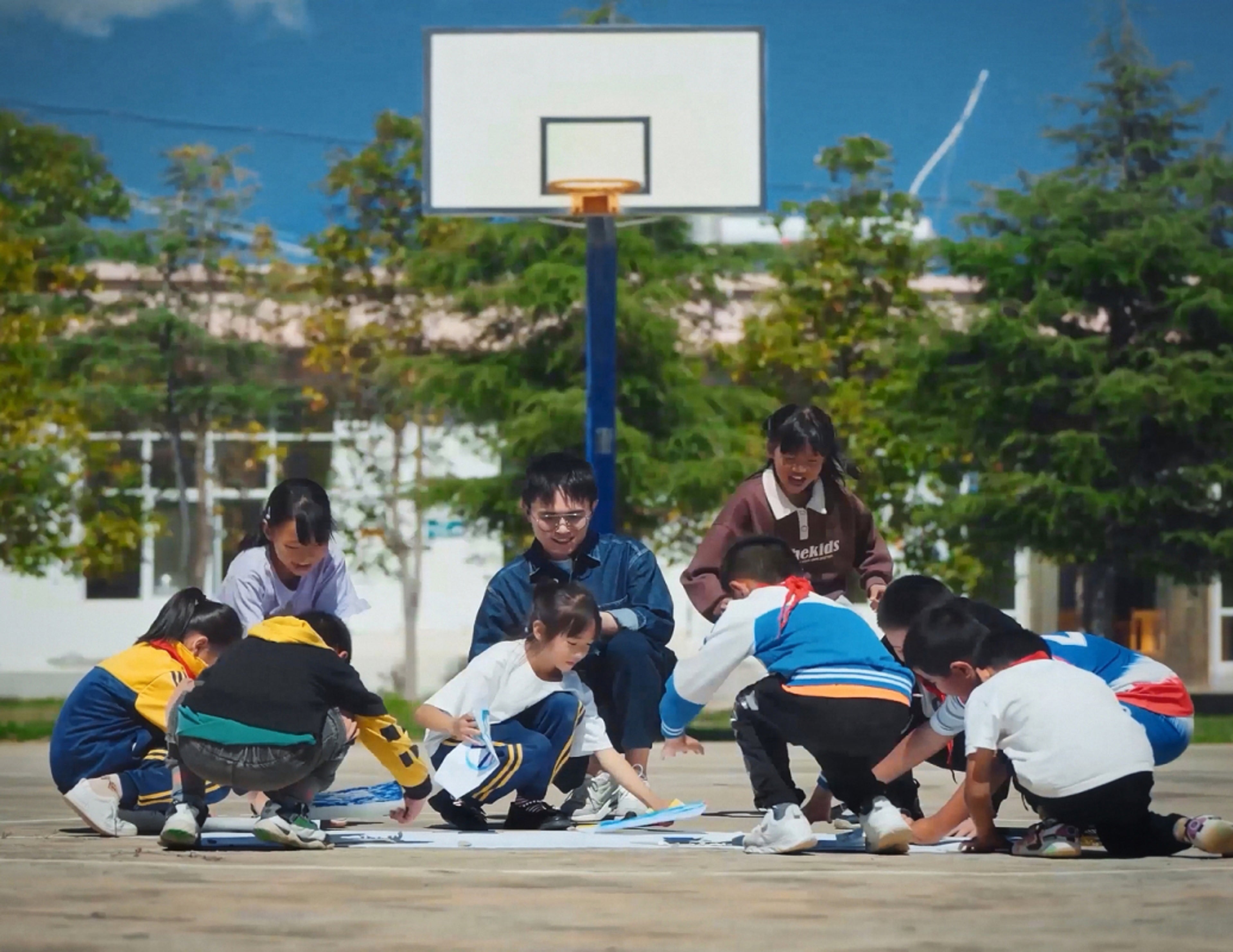 一群亚洲儿童和一位老师在树木环绕的篮球场上展示绘画作品。