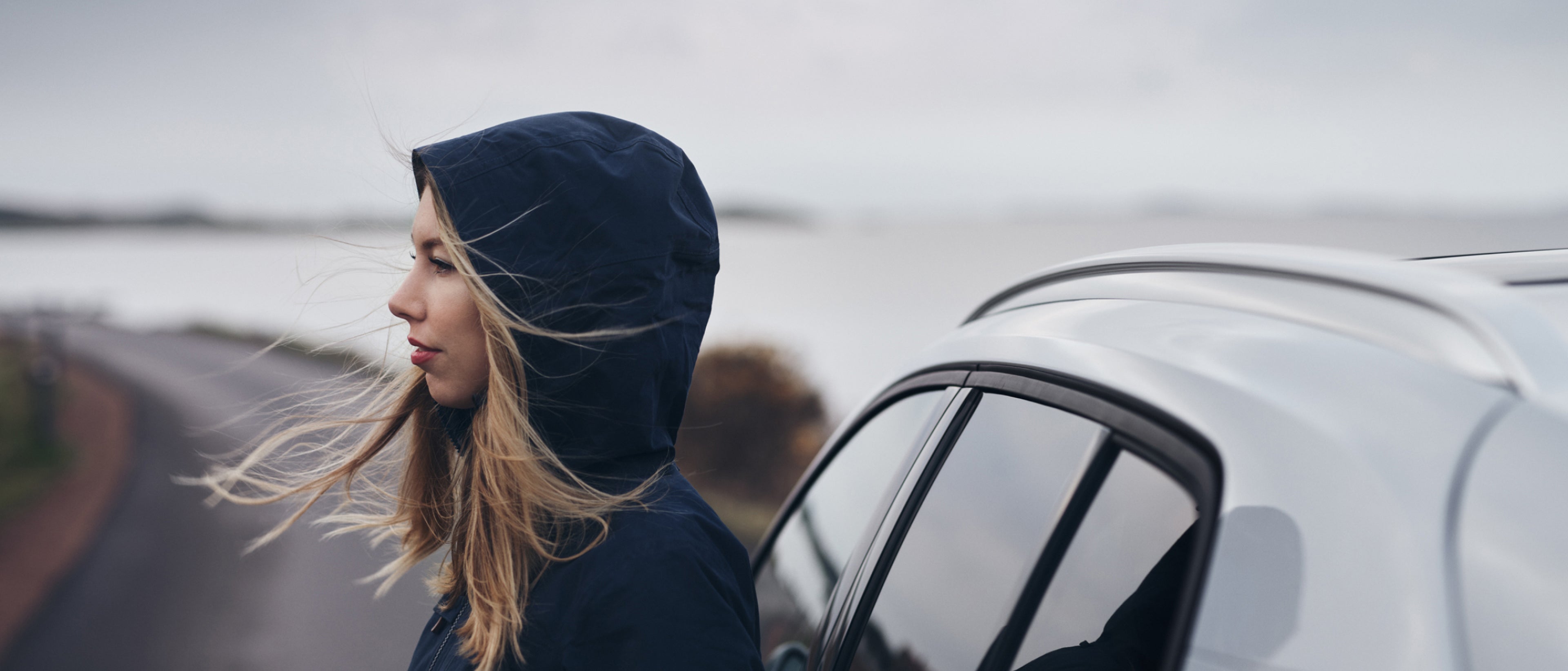 一名女士身穿蓝色连帽夹克,站在白色沃尔沃旁的侧影。