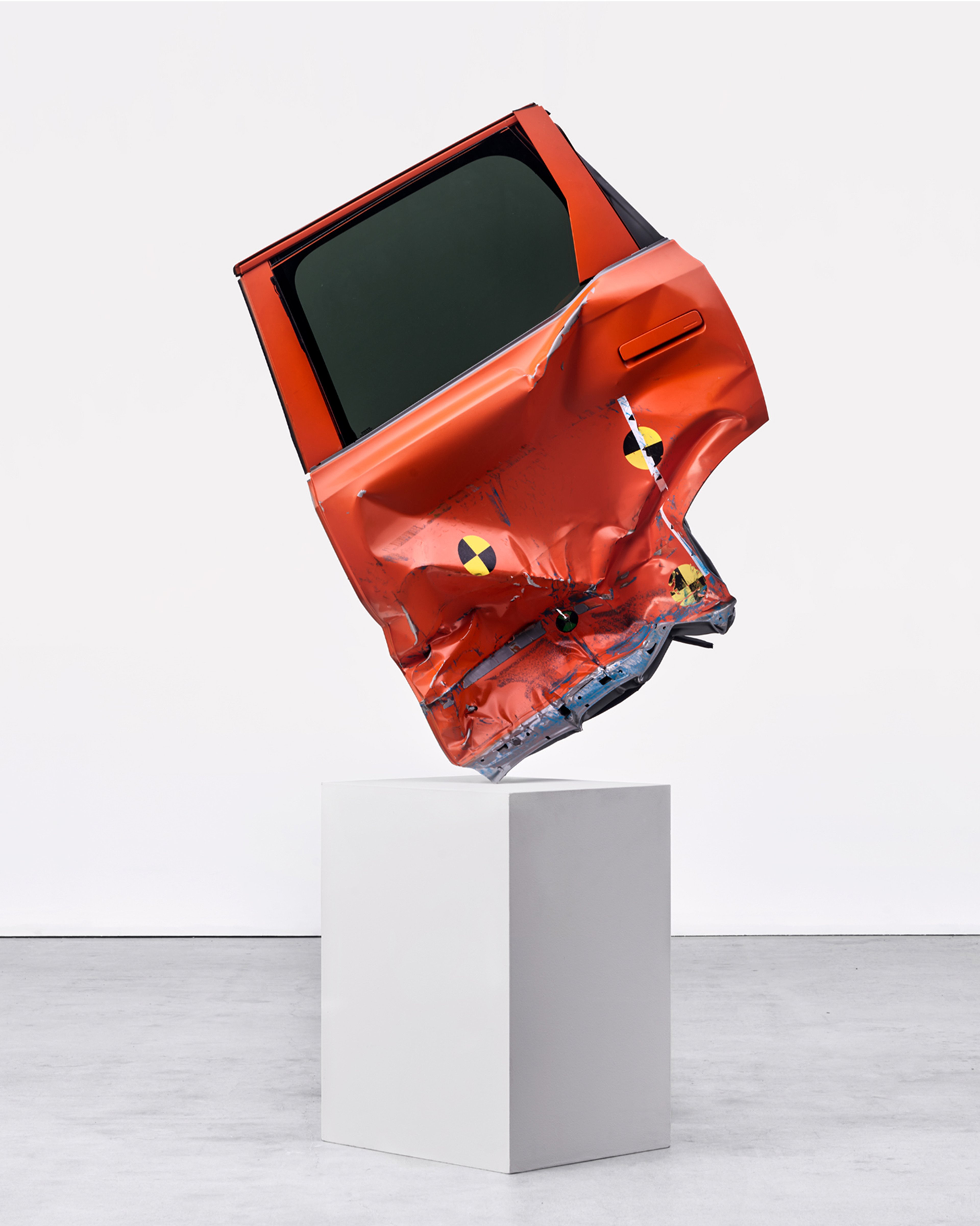 画廊空间的白色基座上直立着一扇压扁的橙色车门,上面有碰撞测试标记。