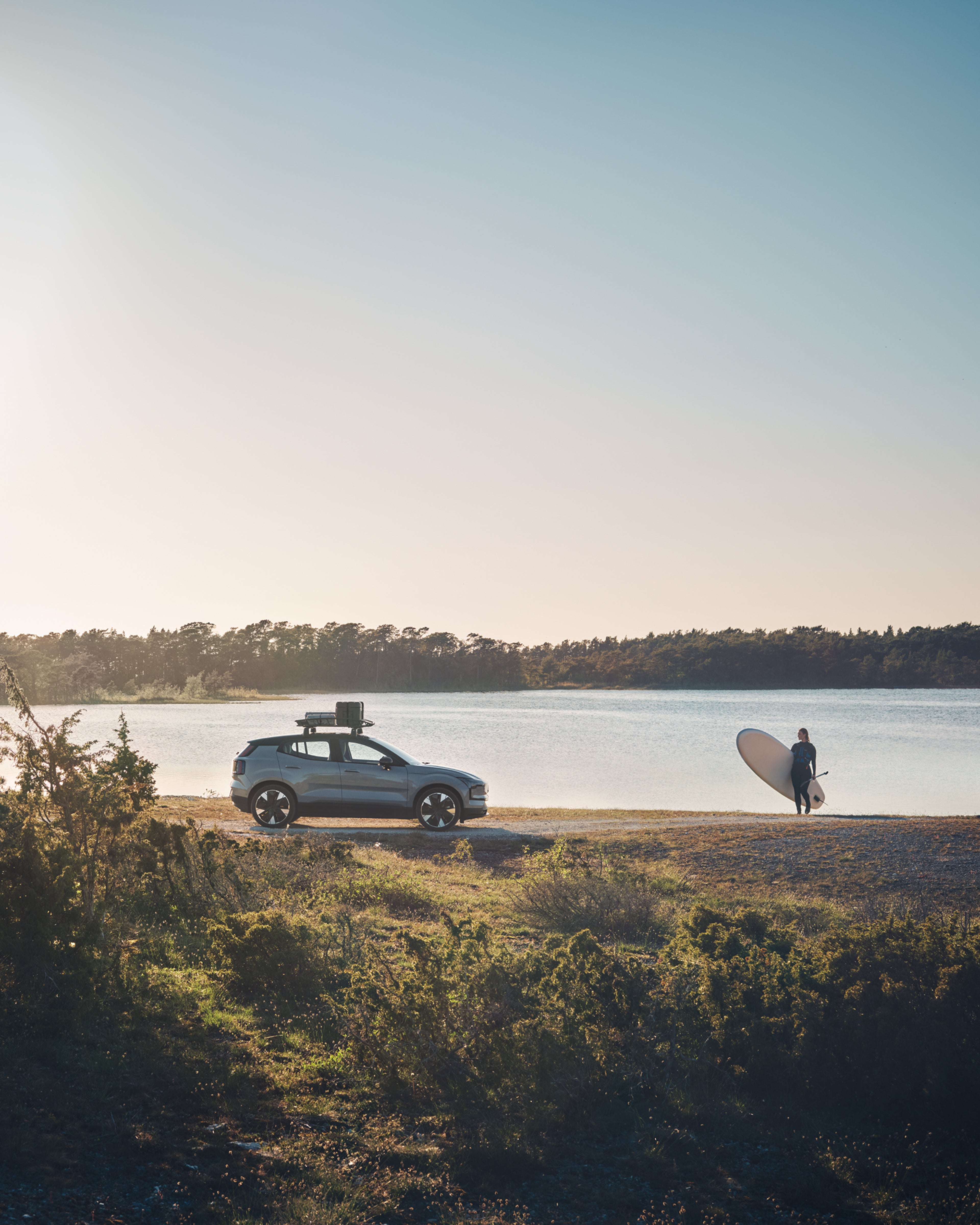 沃尔沃 EX30 侧视图,一位女士拿着冲浪板站在海边。