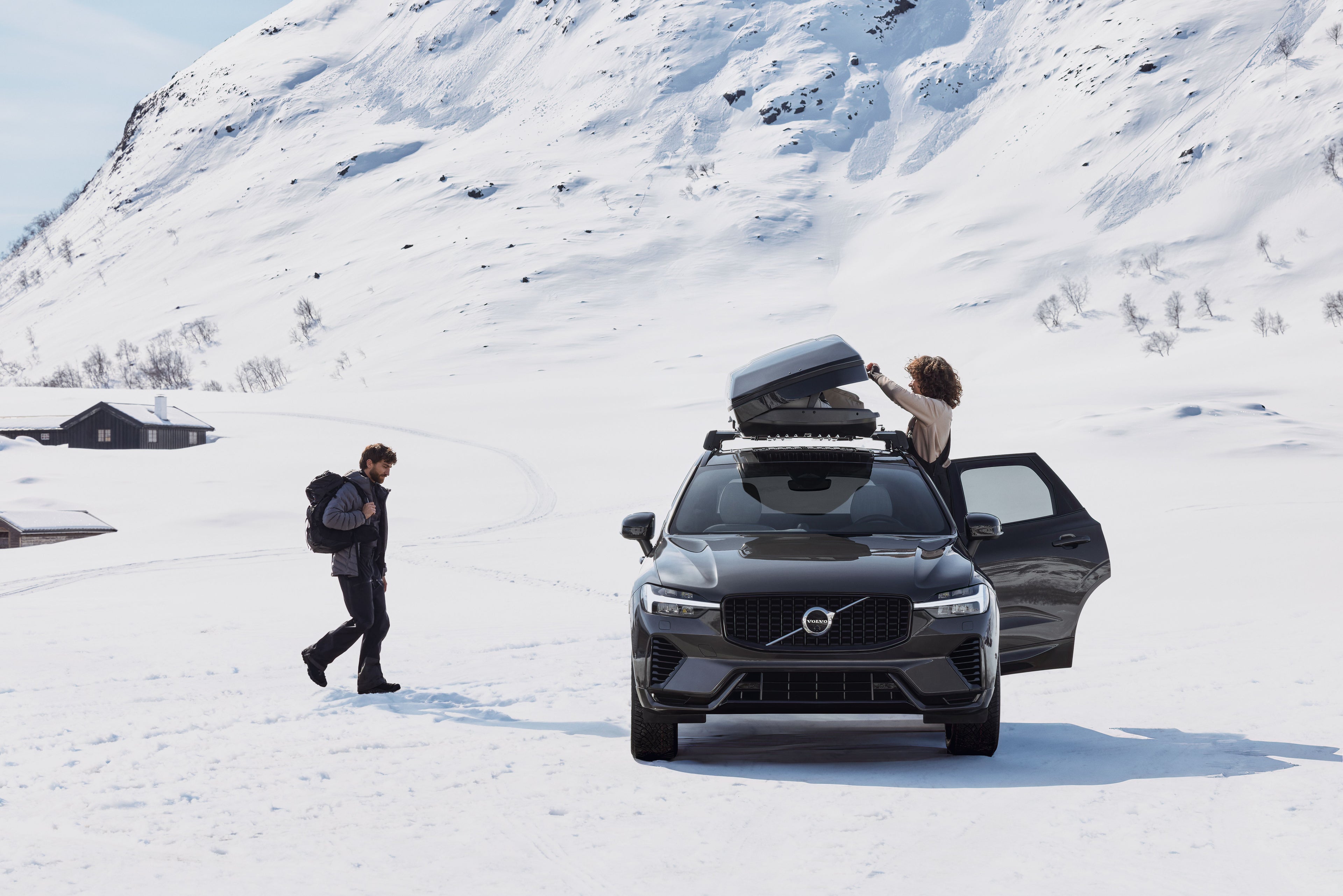 一名女士打开停在雪山附近的一辆沃尔沃 XC60 车顶箱,一位男士走向她的车。