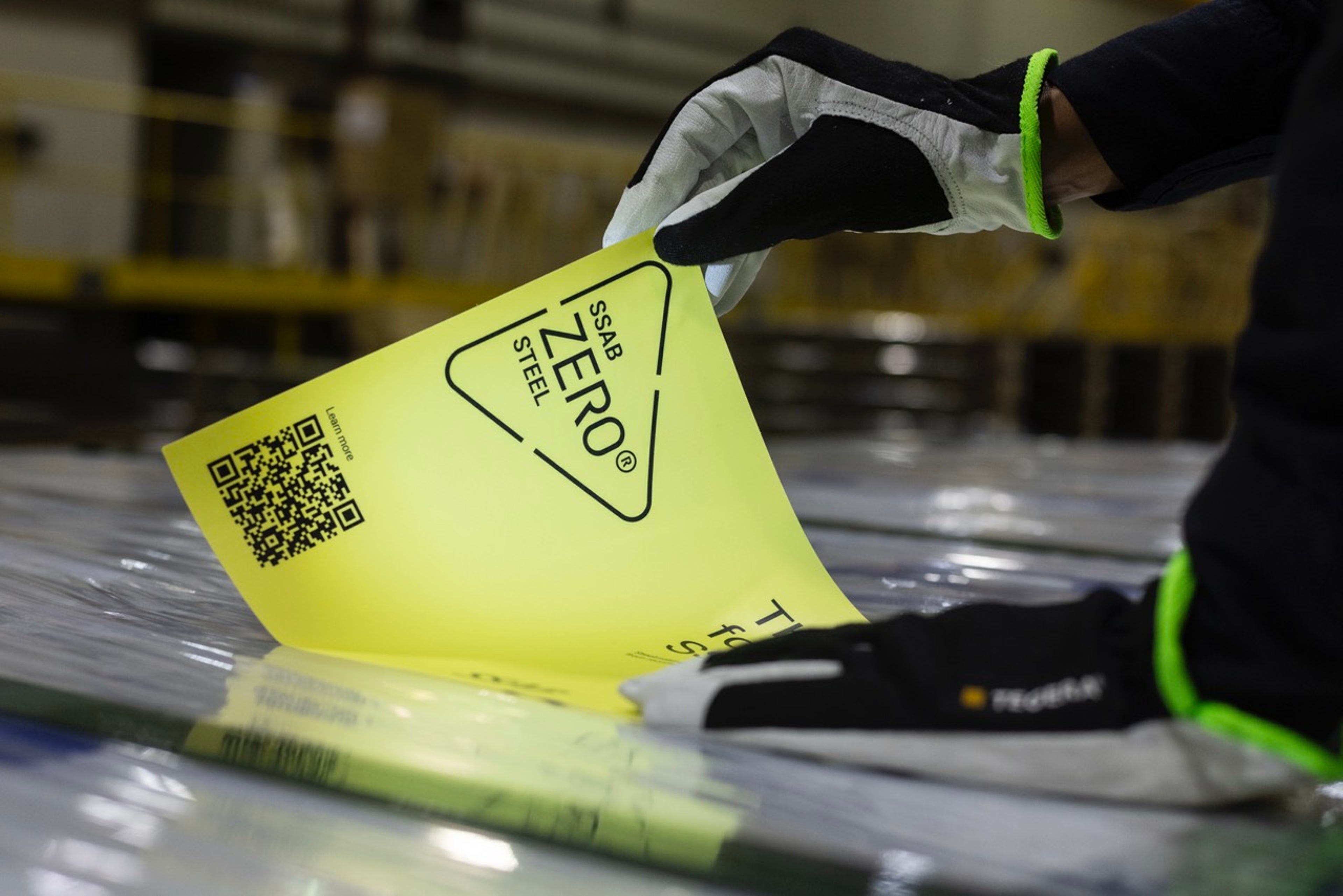一双戴着手套的手揭开印有“SSAB ZERO® STEEL”的黄色标签,露出下面的钢表面。