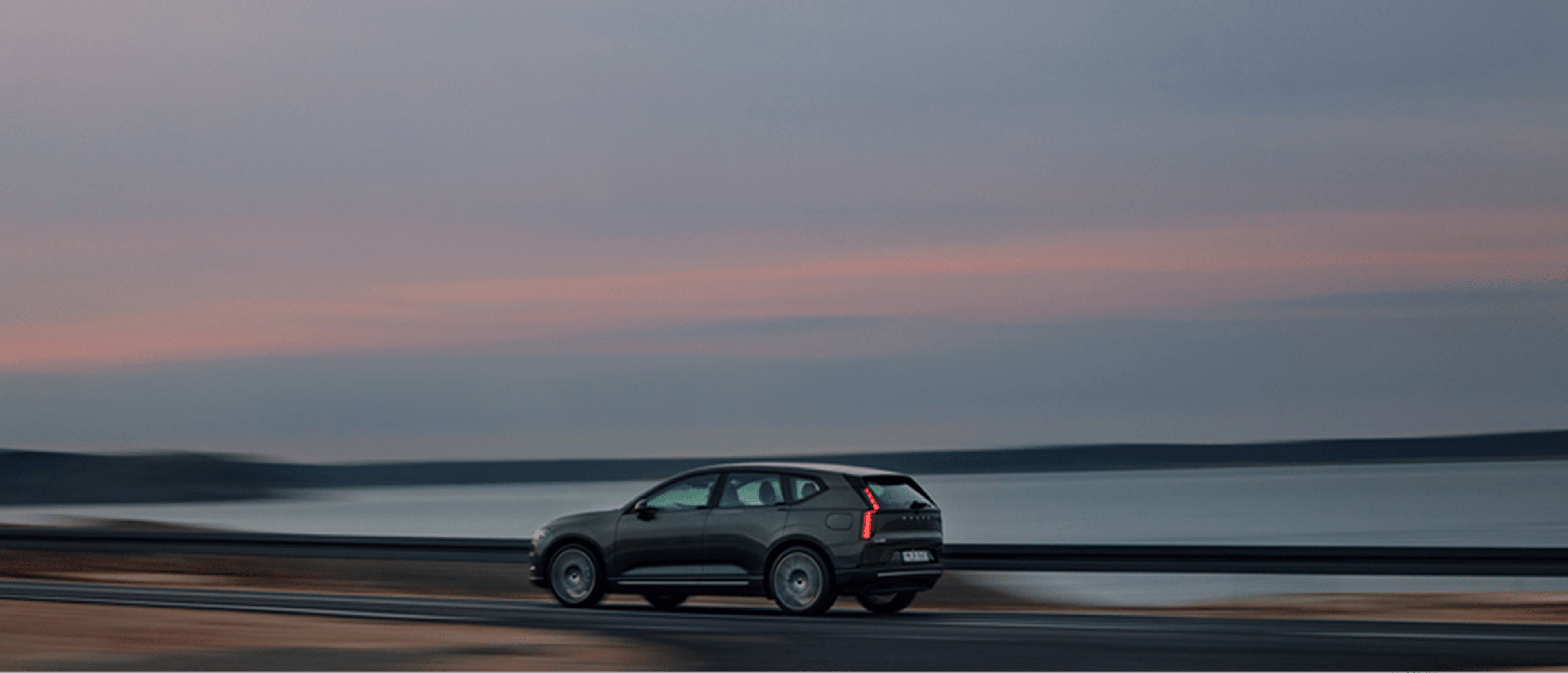 一輛Volvo車沿著靠近水邊的道路行駛,略帶動態模糊以示速度,天空灰藍色,夕陽映照著柔和的粉紅色光澤。