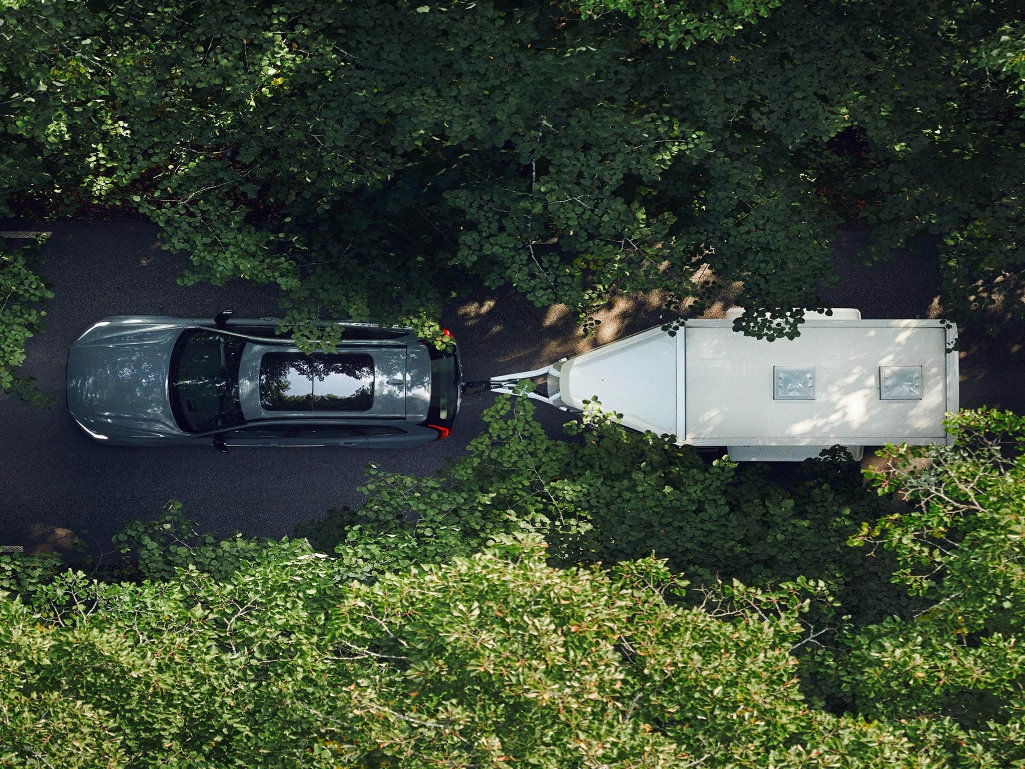 鸟瞰太阳底下,沃尔沃 SUV 沿绿树成荫的道路牵引白色拖车前行。