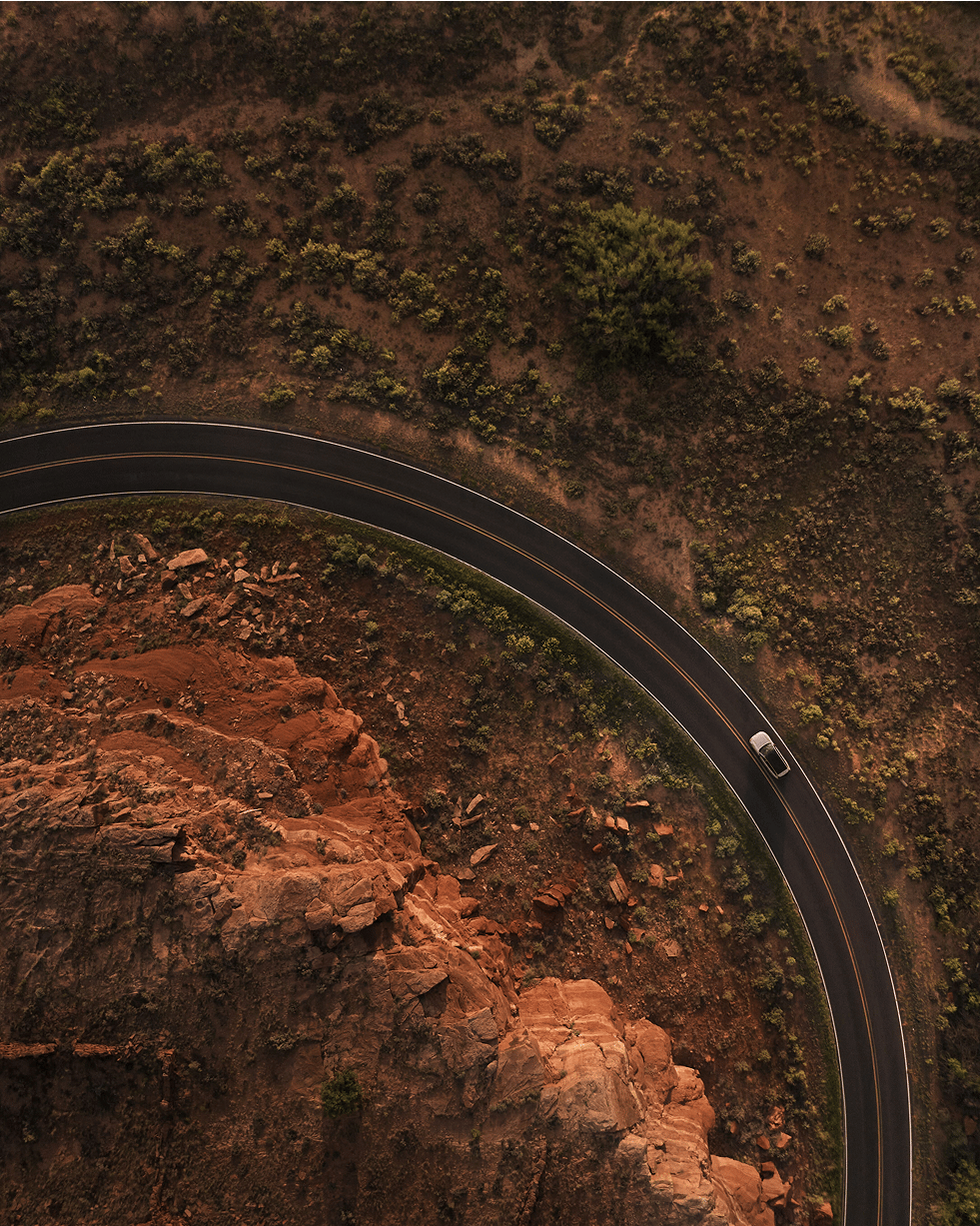 一辆沃尔沃 EX90 沿着一条蜿蜒的道路行驶,这条道路在沙漠景观中急转弯,一侧是红色的岩石峭壁,另一侧是稀疏的植被,从上方捕捉到温暖的金色光线。元件