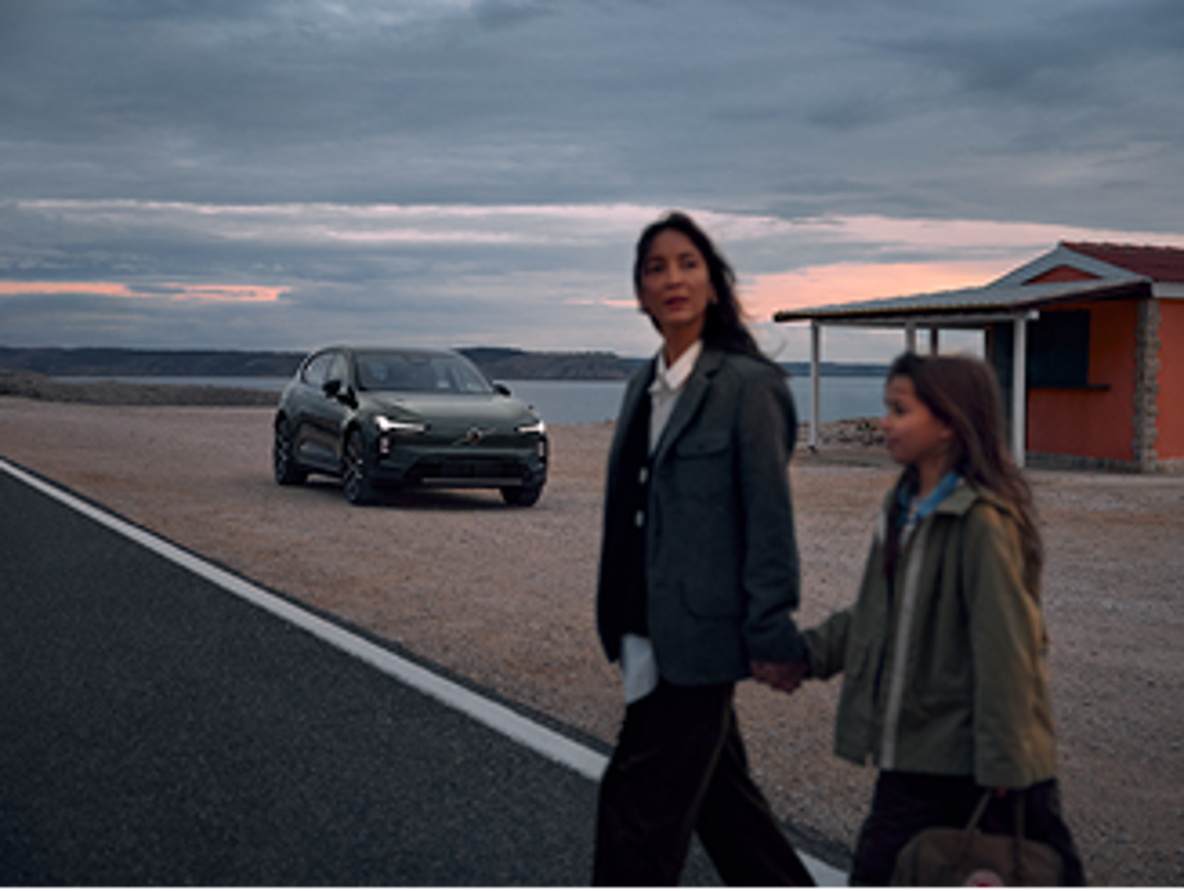 背景中一輛電動車Volvo清晰,前景則是一位模糊的女子手持孩童的手,走過馬路。