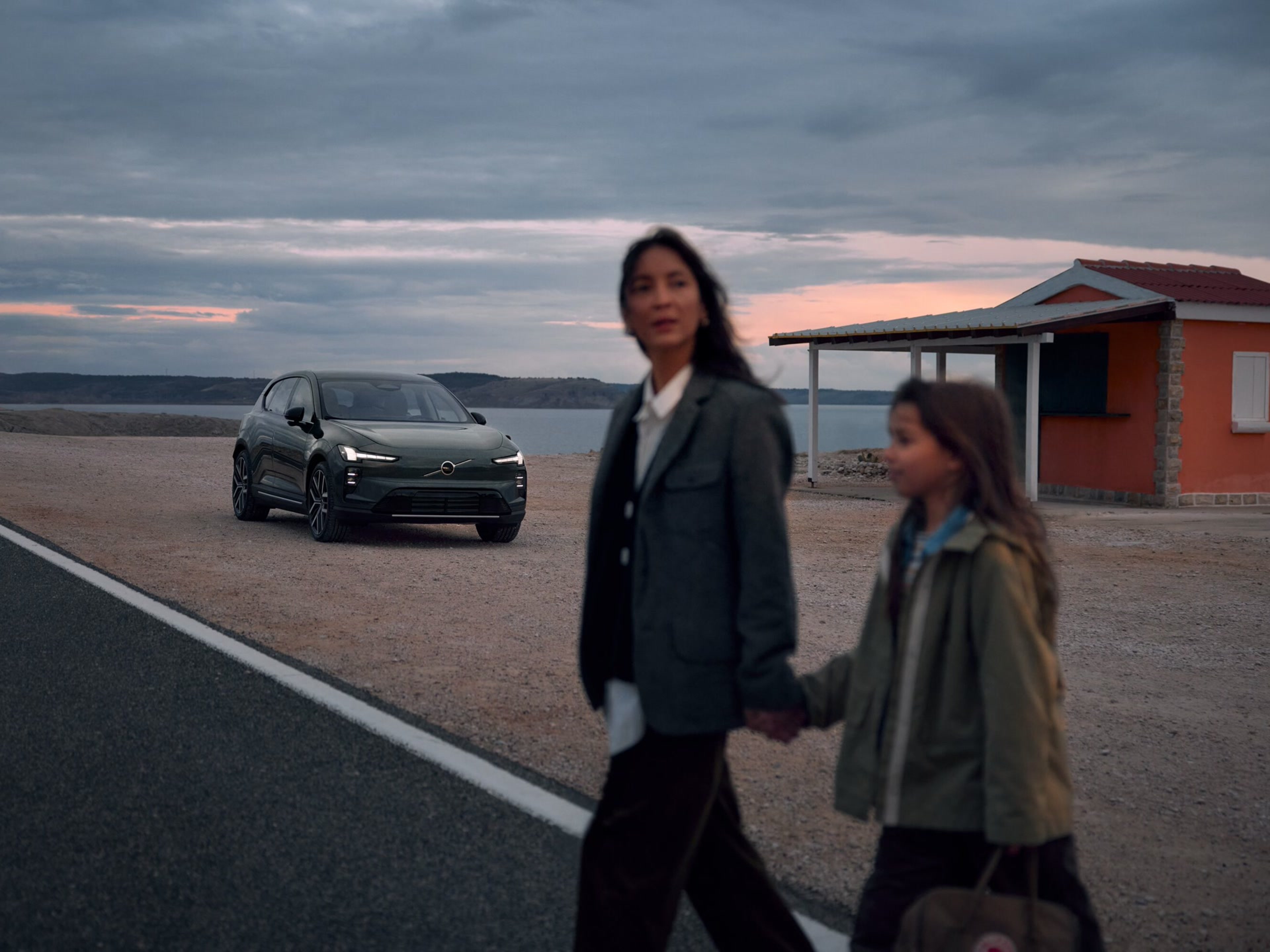 背景中一輛電動車Volvo清晰,前景則是一位模糊的女子手持孩童的手,走過馬路。