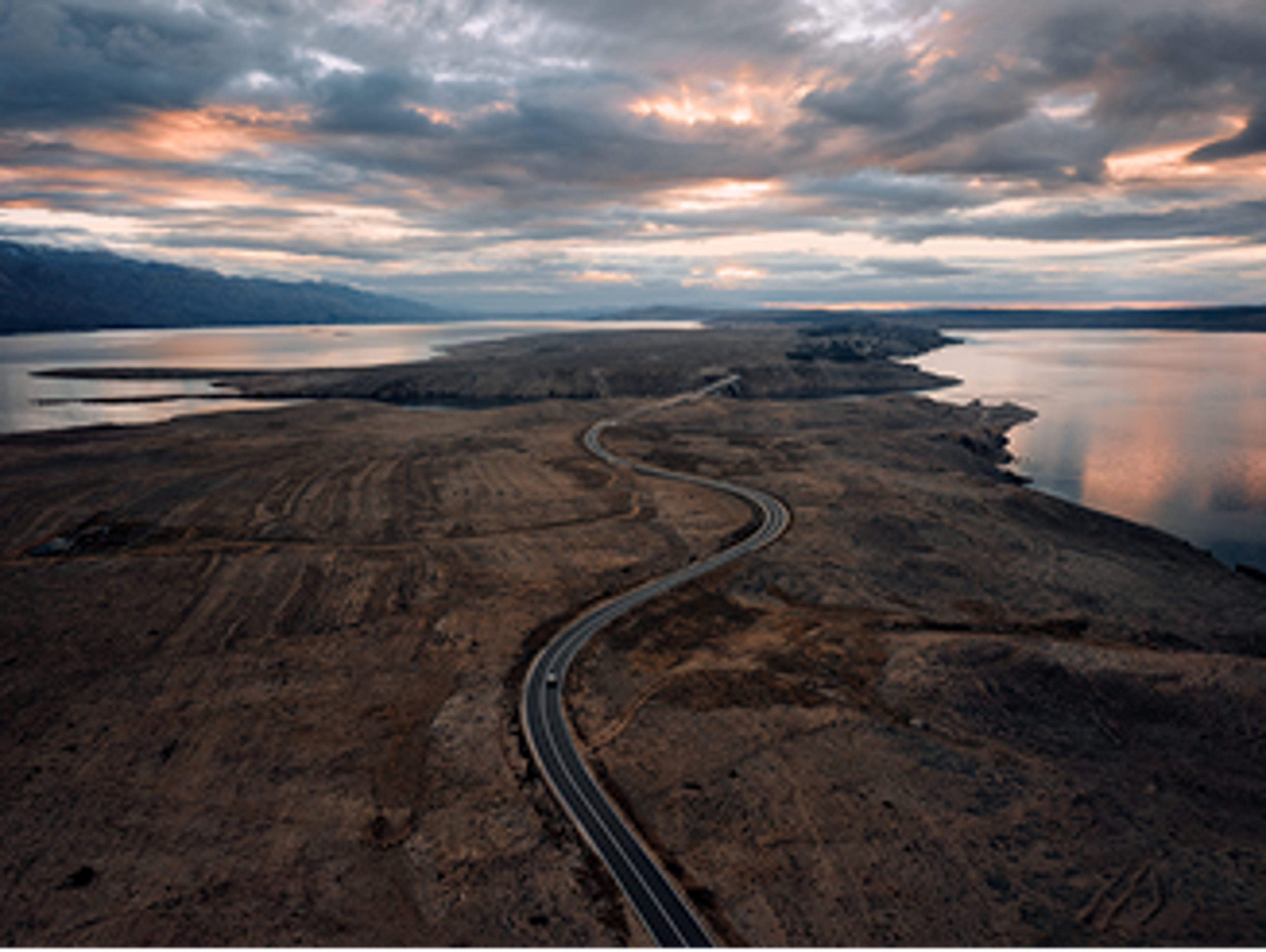 一輛Volvo車沿著蜿蜒陡峭的道路行駛,穿越一片棕色、土色的景觀,兩側的水面映照著陰沉的天空,天空被柔和的粉紅色光芒映照,畫面從上方捕捉到