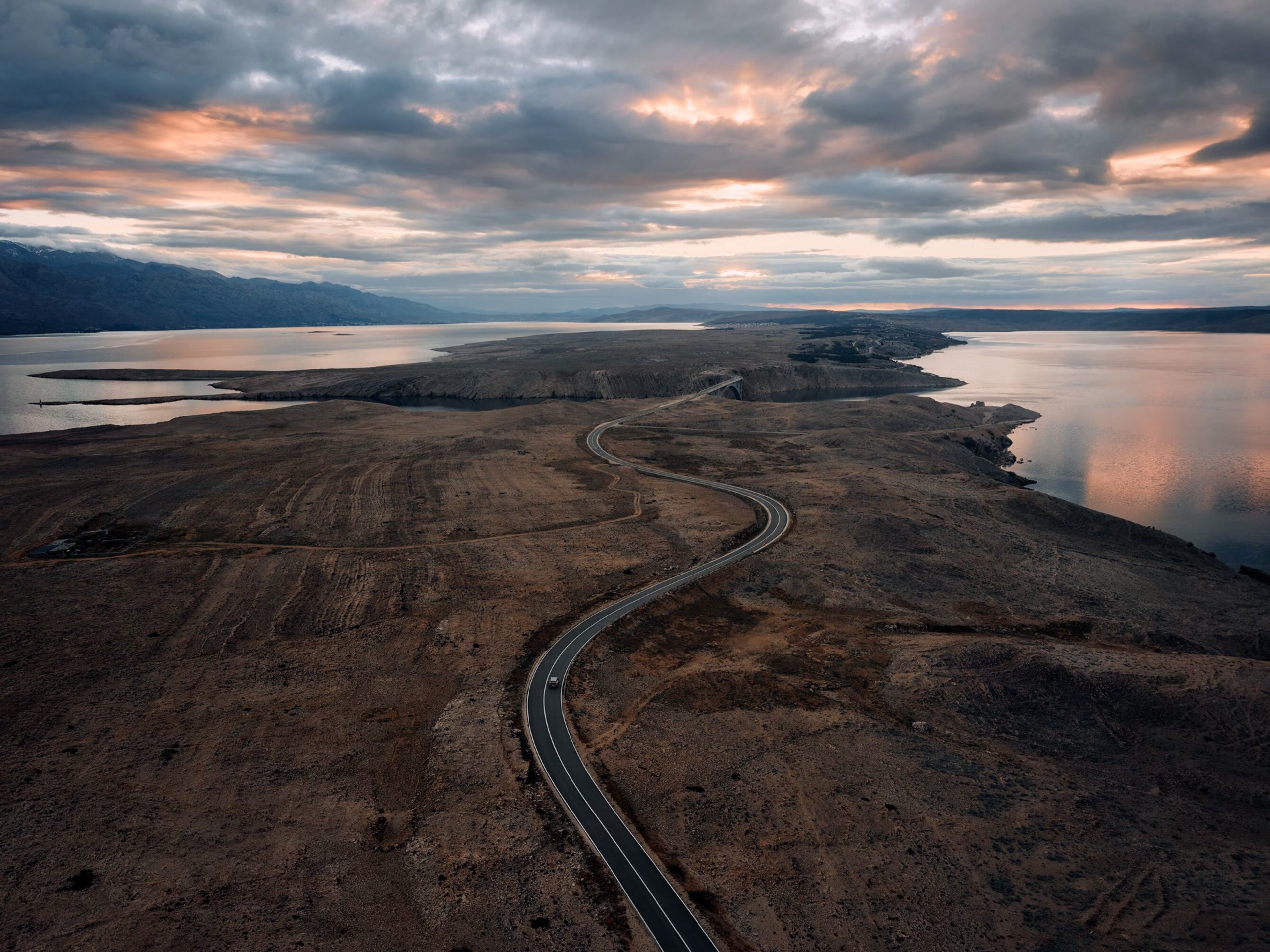 一輛Volvo車沿著蜿蜒陡峭的道路行駛,穿越一片棕色、土色的景觀,兩側的水面映照著陰沉的天空,天空被柔和的粉紅色光芒映照,畫面從上方捕捉到