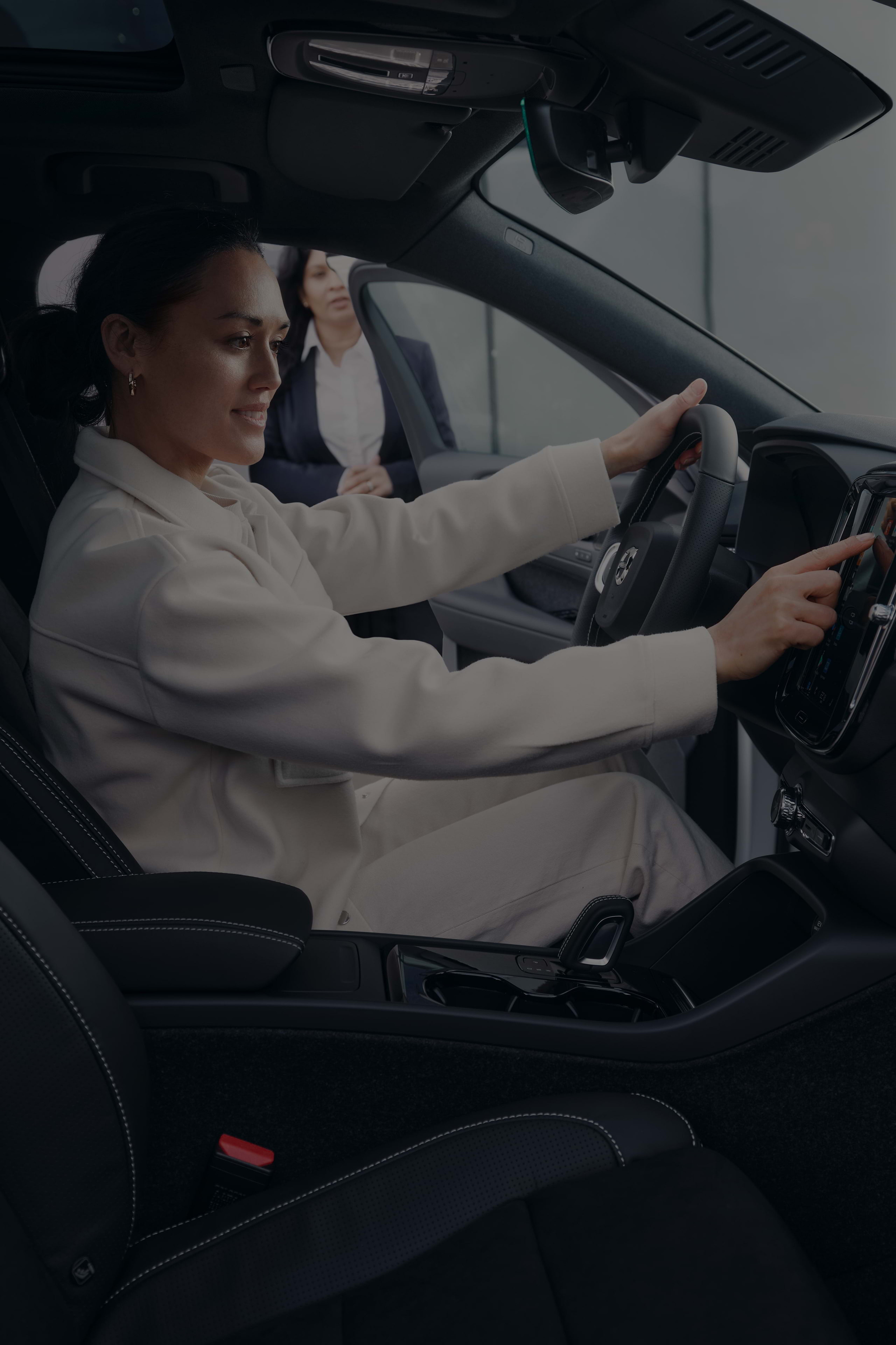 年轻女销售观察另一女性微笑着坐在沃尔沃汽车驾驶员座位上操作中控触摸屏。