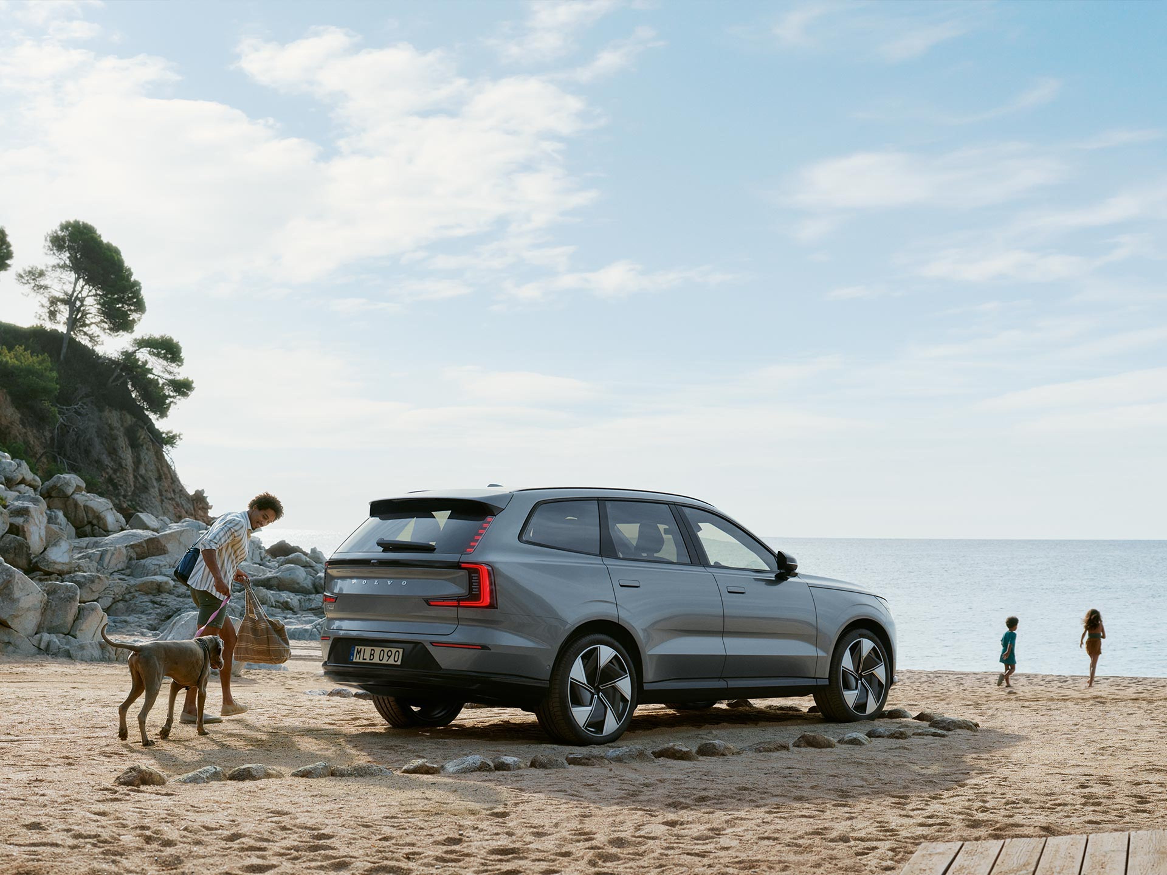 一名男士牵着大狗朝大海走去,孩子们在海边玩耍;家用大型沃尔沃 SUV 停在沙滩上。