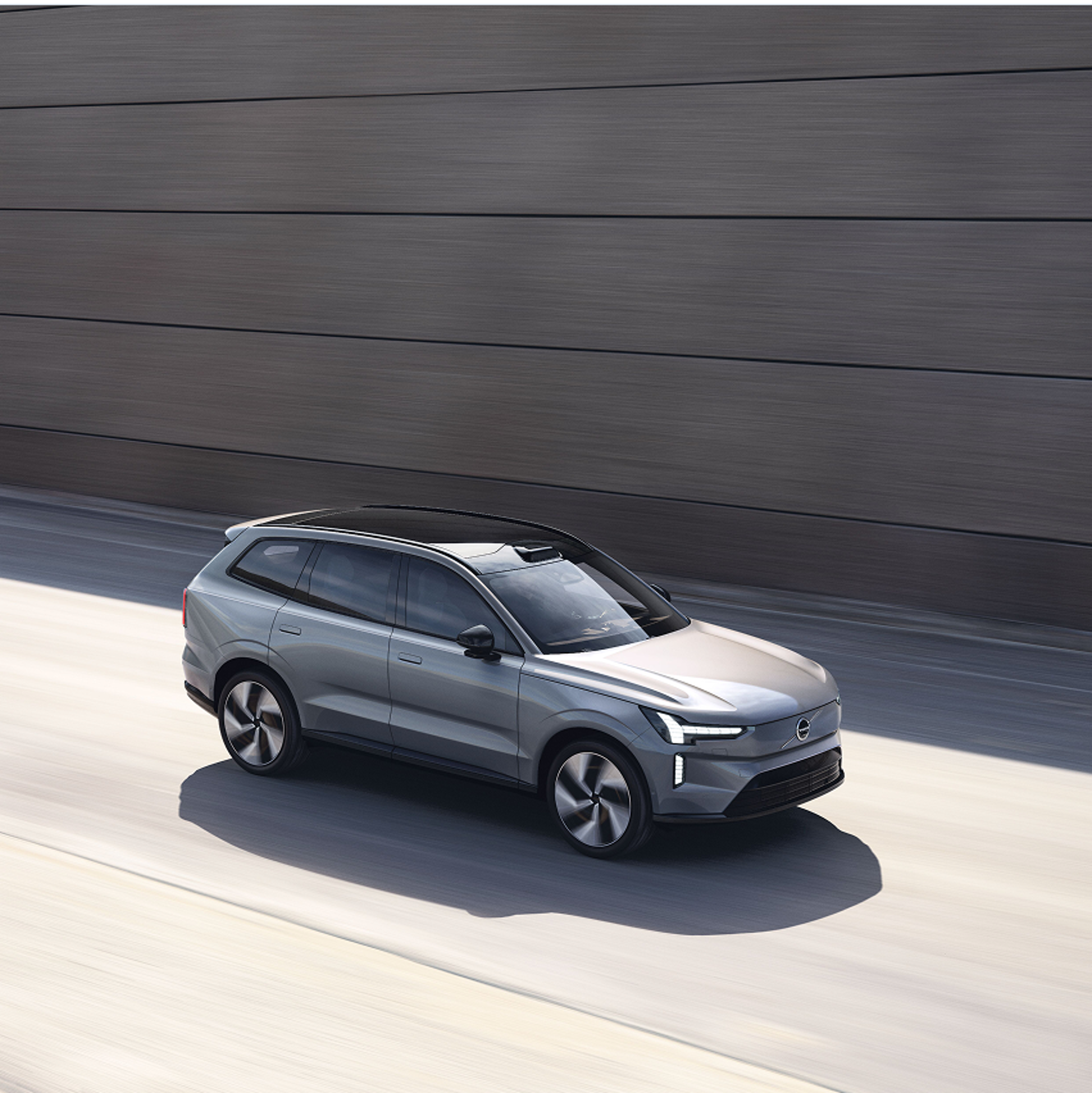 灰色沃尔沃 EX90 行驶在阳光明媚的道路上,旁边是现代混凝土墙。