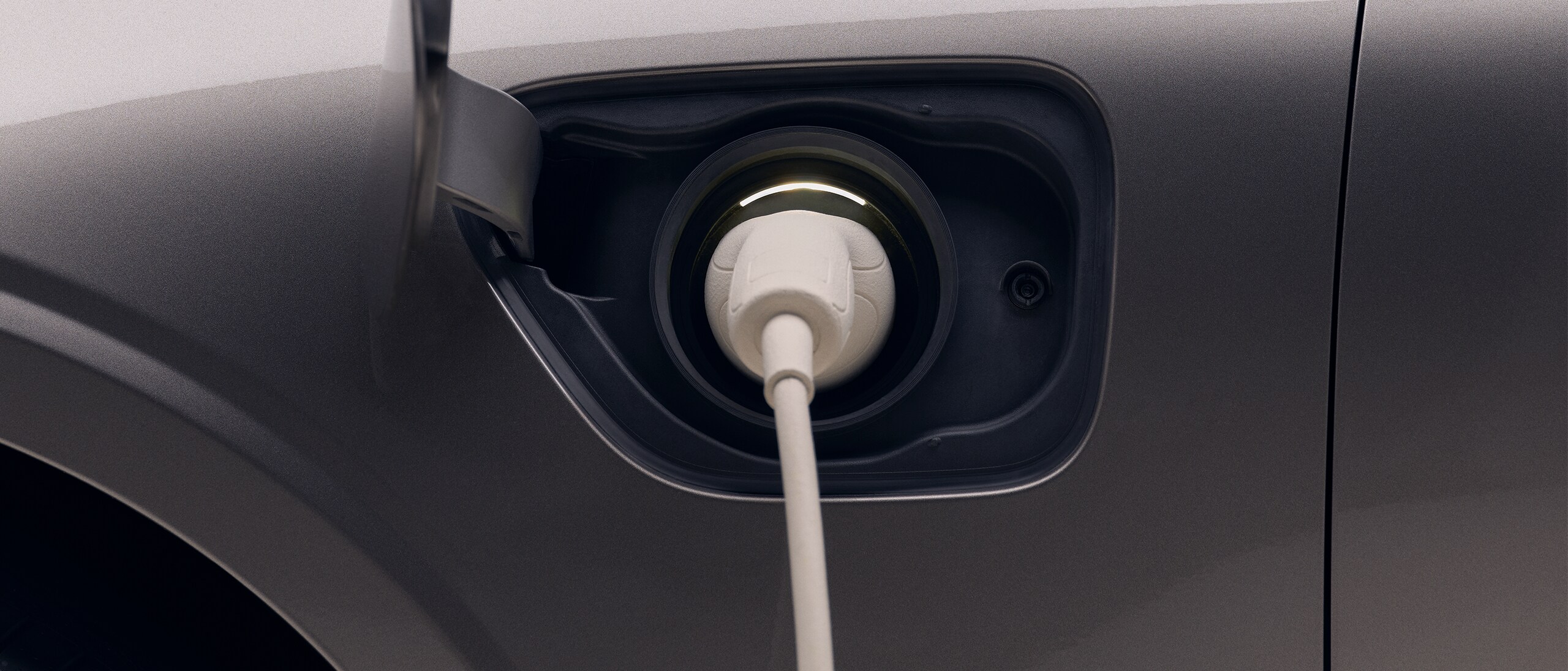 Closeup of the front left quarter of a new dark grey Volvo electric car, with a white charging cable plugged in to the car's charging port.