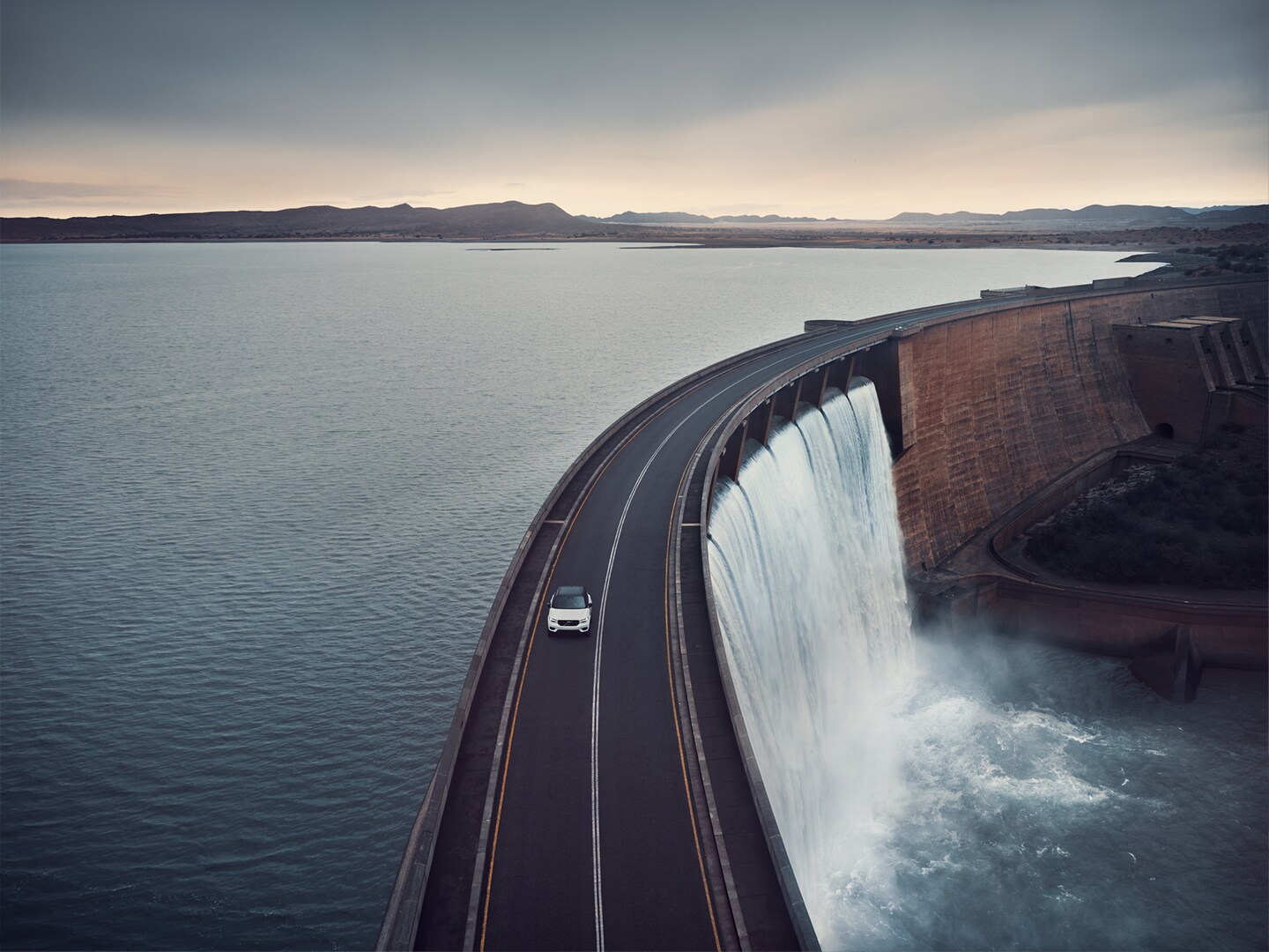 沃尔沃 SUV 在公路上行驶，穿越有水库的大坝。